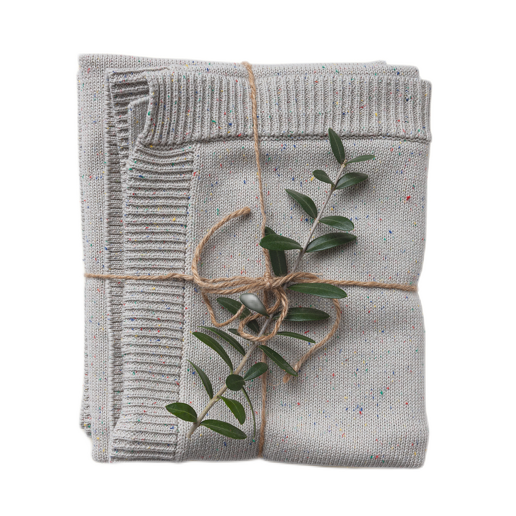 A folded confetti-speckled knit blanket in shades of gray, tied with a string, featuring a small branch of green leaves protruding from the fold.