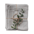 A folded confetti-speckled knit blanket in shades of gray, tied with a string, featuring a small branch of green leaves protruding from the fold.