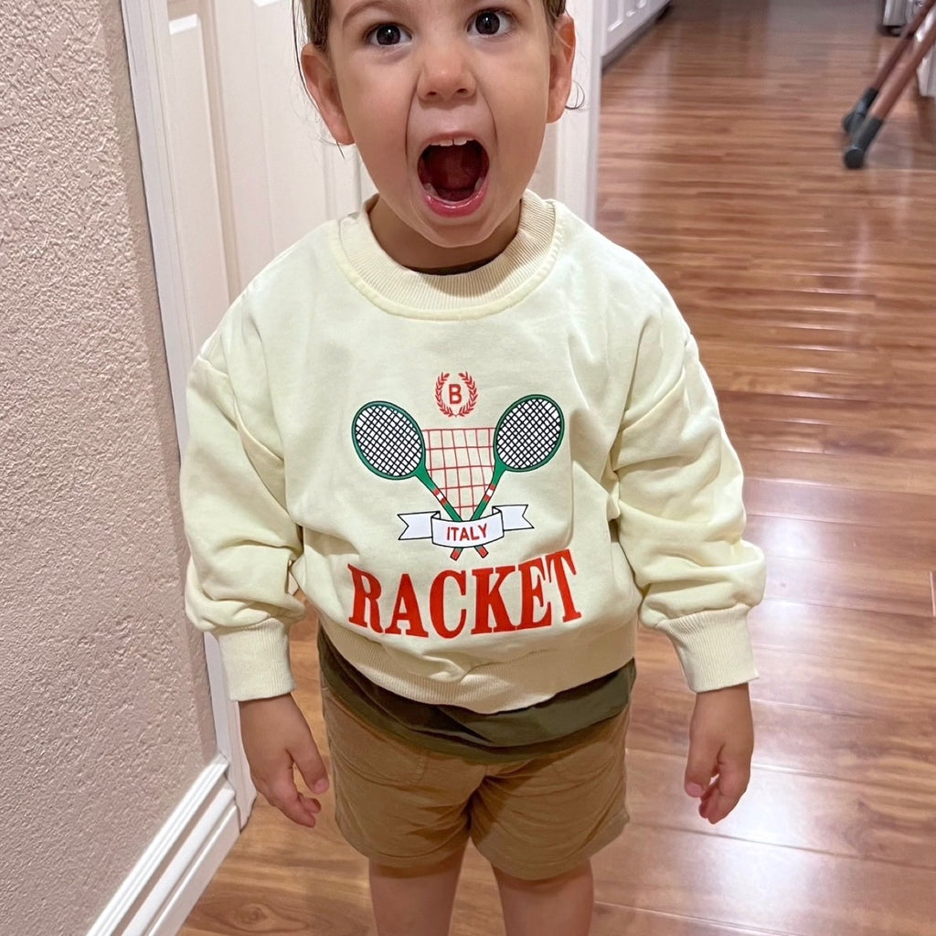 A child wearing a beige sweatshirt with a graphic of a tennis racket and the word 'RACKET' on the front, standing in a home environment.