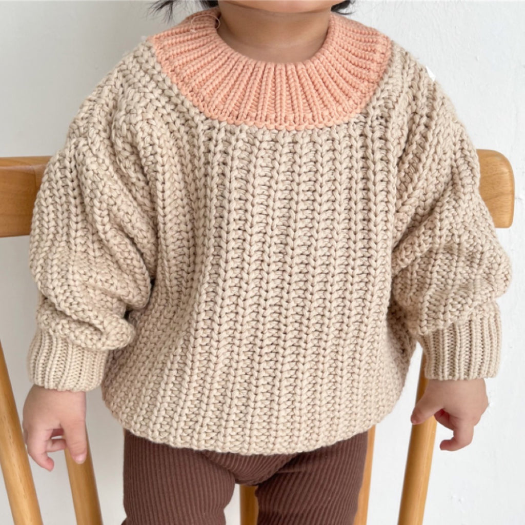A baby wearing a chunky knit sweater with a round neckline and long sleeves, sitting on a wooden chair.